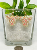 A pair of cow-head dangle earrings with pink and white spotted enameled faces and gold-tone hooks, displayed in a clear glass container filled with white beads, with green leaves above and a coin below for scale.