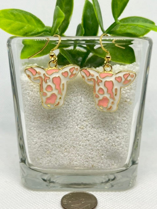 A pair of cow-head dangle earrings with pink and white spotted enameled faces and gold-tone hooks, displayed in a clear glass container filled with white beads, with green leaves above and a coin below for scale.